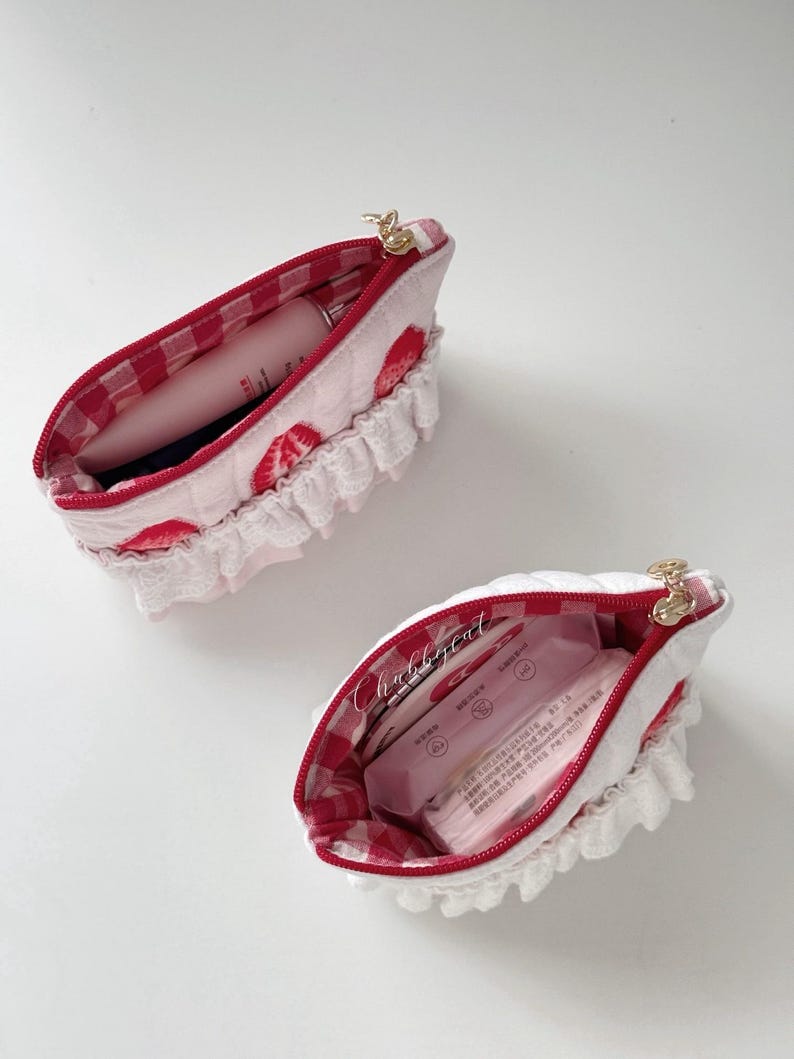 May include: Two quilted pouches featuring a white exterior with red strawberry prints and white ruffled trim. The interiors are lined with red and white gingham fabric. Each pouch has a gold zipper closure and contains various small personal items.