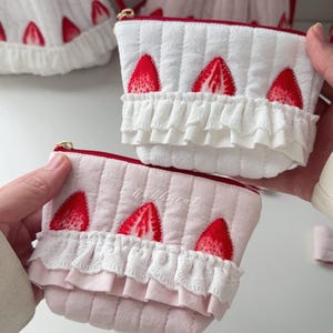 May include: Two quilted pouches featuring a strawberry print and white lace ruffles. One pouch is white and the other is light pink. Both have a red zipper closure at the top. The pink pouch has the word Chubbycat embroidered in white thread.