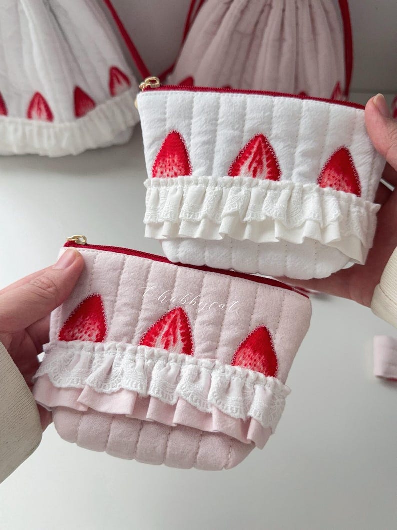 May include: Two quilted pouches featuring a strawberry print and white lace ruffles. One pouch is white and the other is light pink. Both have a red zipper closure at the top. The pink pouch has the word Chubbycat embroidered in white thread.