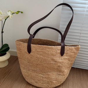 May include: A woven straw tote bag with dark brown leather handles. The bag is a light tan color and has a rounded shape. The handles are attached to the bag with dark brown leather straps.