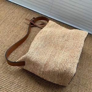 May include: A woven straw shoulder bag with a brown leather strap. The bag is rectangular with a rounded top and a brown leather strap. The bag is made of a light tan woven material.
