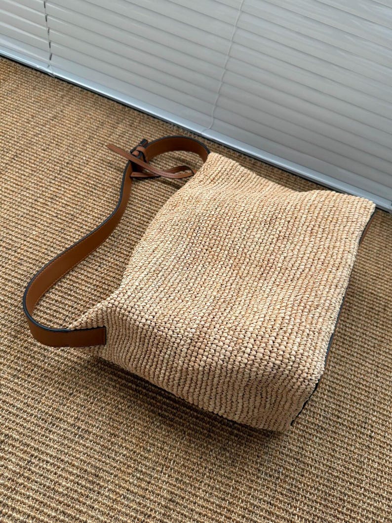 May include: A woven straw shoulder bag with a brown leather strap. The bag is rectangular with a rounded top and a brown leather strap. The bag is made of a light tan woven material.
