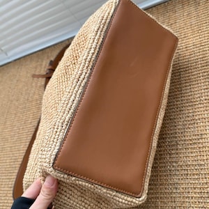 May include: A woven straw tote bag with a brown leather base and trim. The bag has a shoulder strap. The bag is a light tan colour. The bag is held by a person's hand.