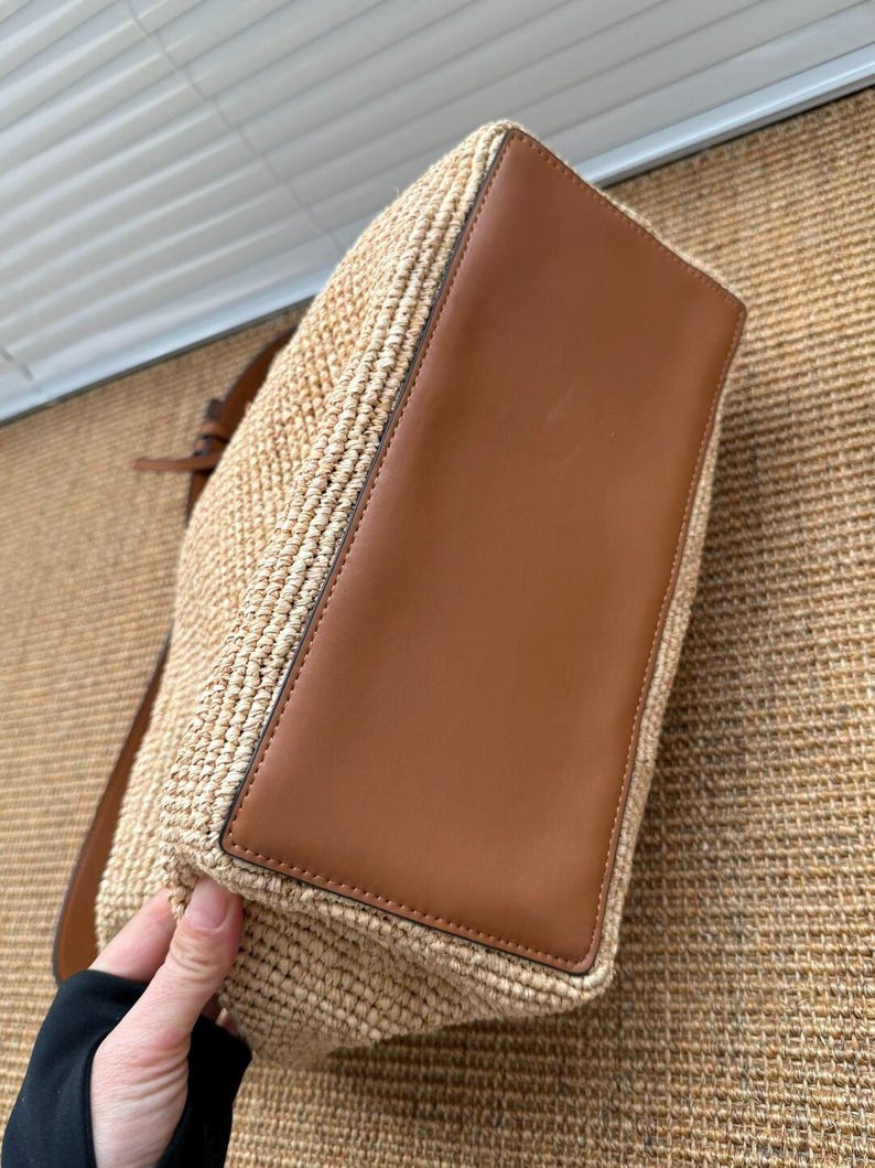 May include: A woven straw tote bag with a brown leather base and trim. The bag has a shoulder strap. The bag is a light tan colour. The bag is held by a person's hand.