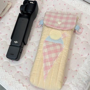 Könnte beinhalten: Eine schwarze DJI Handheld-Gimbal-Kamera neben einer gesteppten Stofftasche mit einer rosa-weiß karierten Klappe, einem hellgelben Körper und einer Eiswaffel-Applikation mit kleinem weißem Herzknopfverschluss und hellblauen Seitenringen.