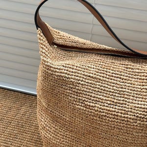 May include: A beige woven raffia handbag with a brown leather handle and a black zipper. The bag has a natural colour and appears handmade. The handle is curved and attached to the top of the bag.