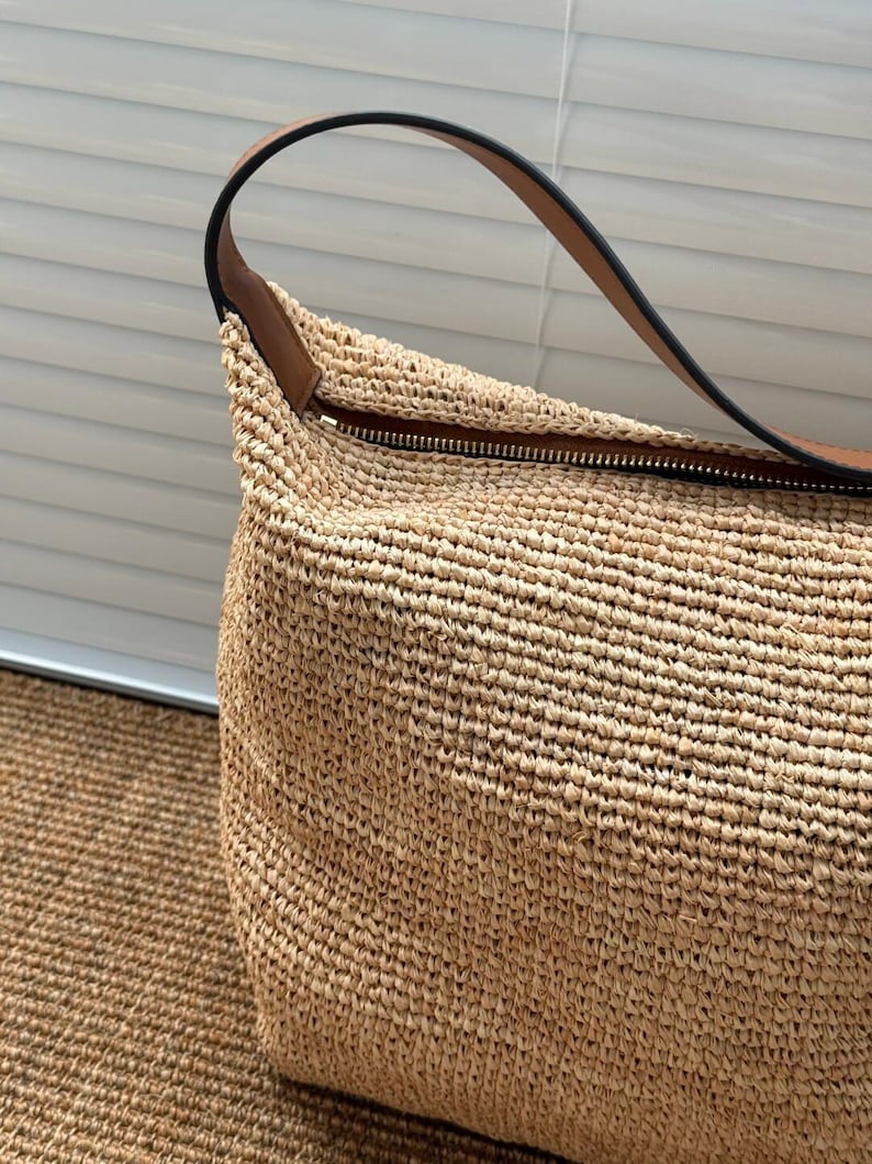 May include: A beige woven raffia handbag with a brown leather handle and a black zipper. The bag has a natural colour and appears handmade. The handle is curved and attached to the top of the bag.