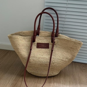 May include: A beige woven straw tote bag with brown leather handles and a long shoulder strap. The bag features a rectangular leather patch with text.