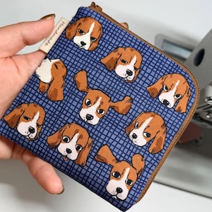 May include: A square zippered pouch featuring a repeating pattern of brown and white beagle dog faces on a blue grid background. The pouch has a brown zipper and a small white fabric tag on the side with the word Handmade printed in black text.