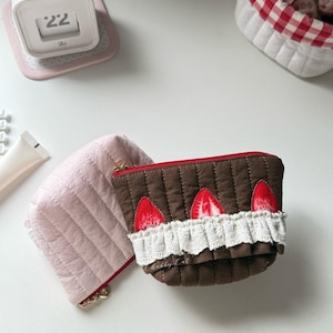 May include: Two quilted zippered pouches. One is solid light pink. The other is brown with a red zipper, featuring three red strawberry appliques and a white ruffled lace trim along the front. The brown pouch has the text Chubby Cat printed on it.