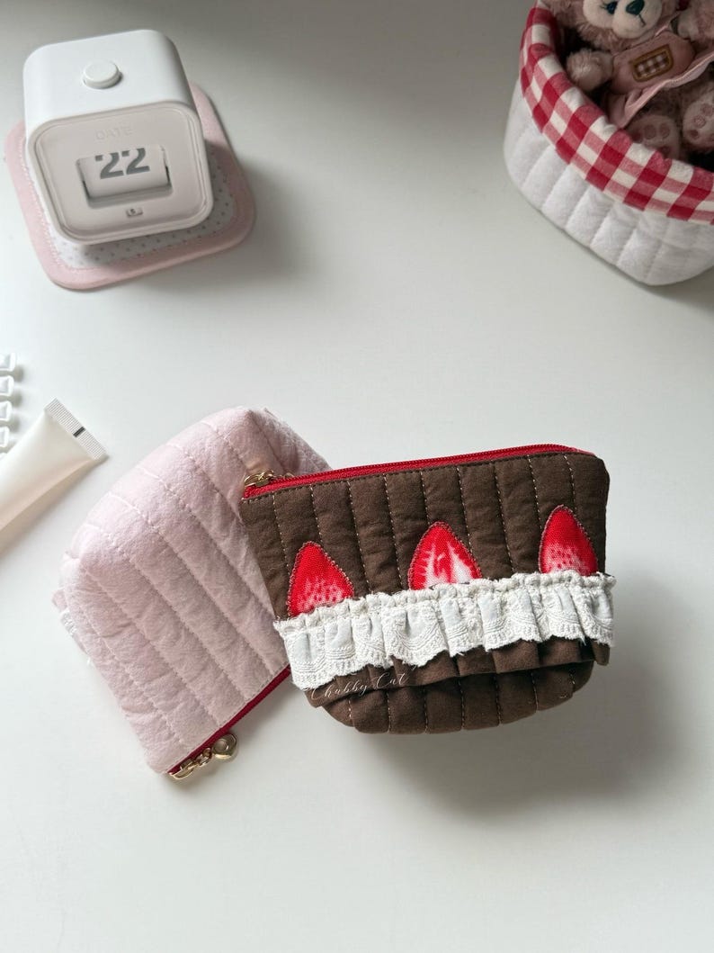 May include: Two quilted zippered pouches. One is solid light pink. The other is brown with a red zipper, featuring three red strawberry appliques and a white ruffled lace trim along the front. The brown pouch has the text Chubby Cat printed on it.