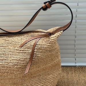 May include: A woven straw shoulder bag with a brown leather strap and zip. The bag is a natural beige colour with a textured surface. The strap is attached with a brown leather loop.