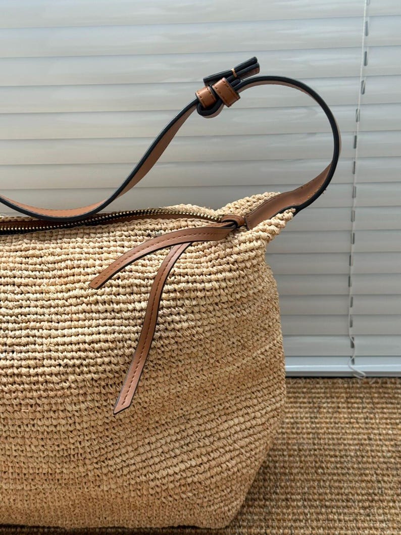 May include: A woven straw shoulder bag with a brown leather strap and zip. The bag is a natural beige colour with a textured surface. The strap is attached with a brown leather loop.