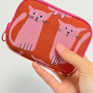 May include: A rectangular fabric zipper pouch featuring a repeating pattern of pink cats on a burnt orange background. The pouch has a bright pink zipper closure along the top and sides, with a matching pink circular zipper pull attached to the slider.