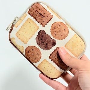 May include: A rectangular zippered pouch featuring a cream-colored fabric printed with various brown and tan cookies, including a bear-shaped cookie and a rectangular cookie with the word YUMMY printed on it, finished with a beige zipper.