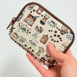 May include: A small, rectangular, beige zippered pouch featuring a repeating pattern of coffee-themed illustrations, including coffee beans, cups, a French press, a bag labeled COFFEE BEANS, and the text Café and Coffee in a dark brown font.