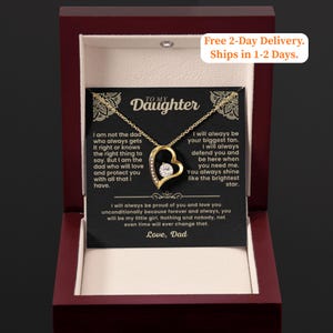 Op de afbeelding: Een goudkleurige hartvormige hangerketting met een heldere steen in het midden, gepresenteerd in een donkerrode sieradendoos. De doos bevat een kaart met de tekst "To My Daughter" en een bericht van "Love, Dad."