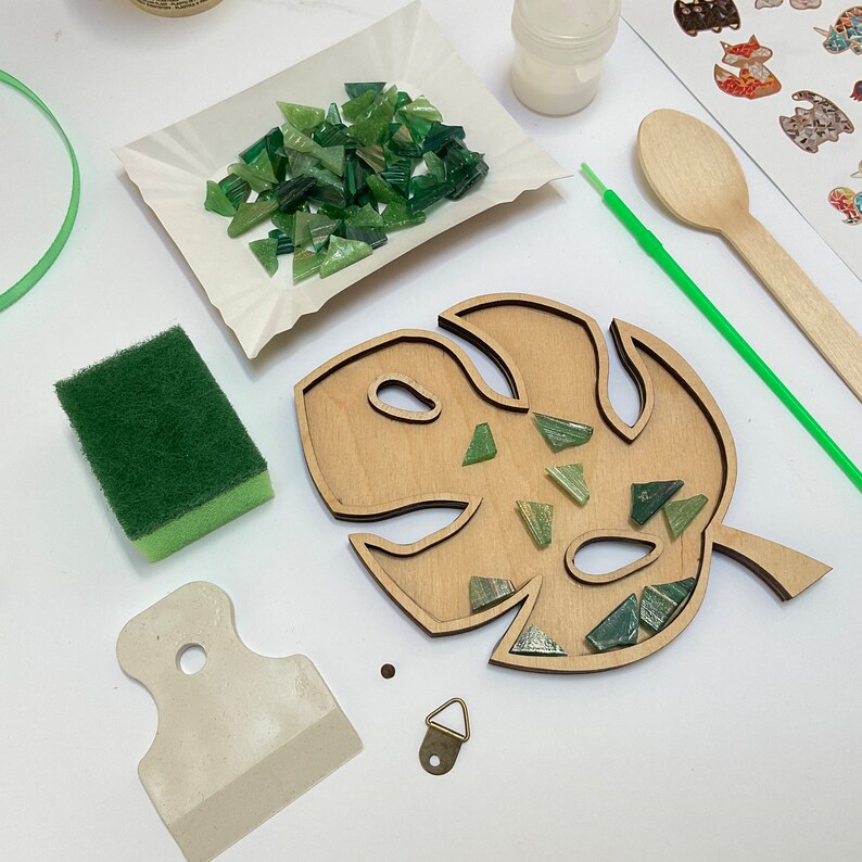 May include: A DIY mosaic kit featuring a wooden monstera leaf with a cutout design. The kit includes green glass tiles, a scraper, a sponge, and a hanging hook.