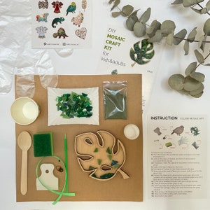 May include: DIY mosaic craft kit for kids and adults. The kit includes a wooden monstera leaf, green glass tiles, green grout, a wooden stick, a sponge, a small cup, a plastic bag, and instructions.