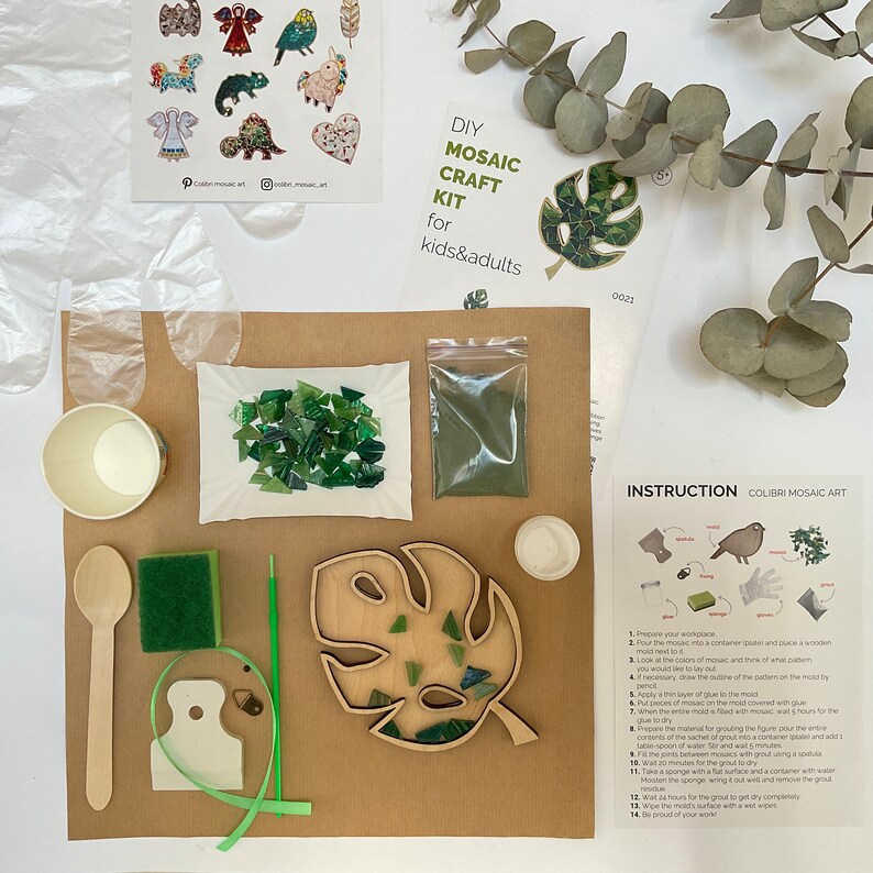 May include: DIY mosaic craft kit for kids and adults. The kit includes a wooden monstera leaf, green glass tiles, green grout, a wooden stick, a sponge, a small cup, a plastic bag, and instructions.