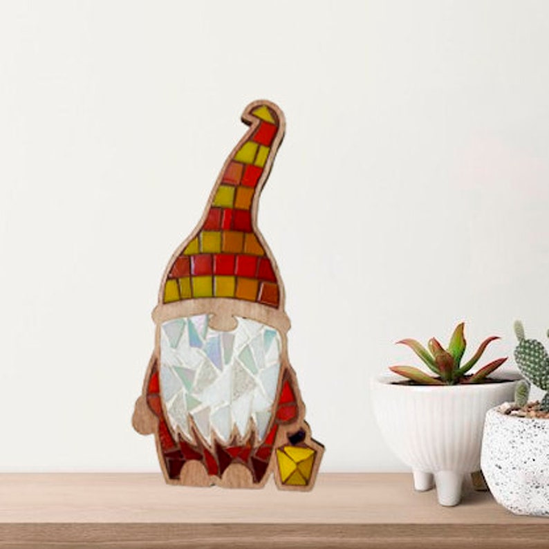 Puede incluir: Figura de gnomo de madera con dise&ntilde;o de mosaico. El gnomo tiene un sombrero colorido con cuadrados rojos, naranjas y amarillos. La barba est&aacute; hecha de piezas de vidrio blancas e iridiscentes. Sostiene una linterna amarilla. La figura est&aacute; sobre una superficie de madera.