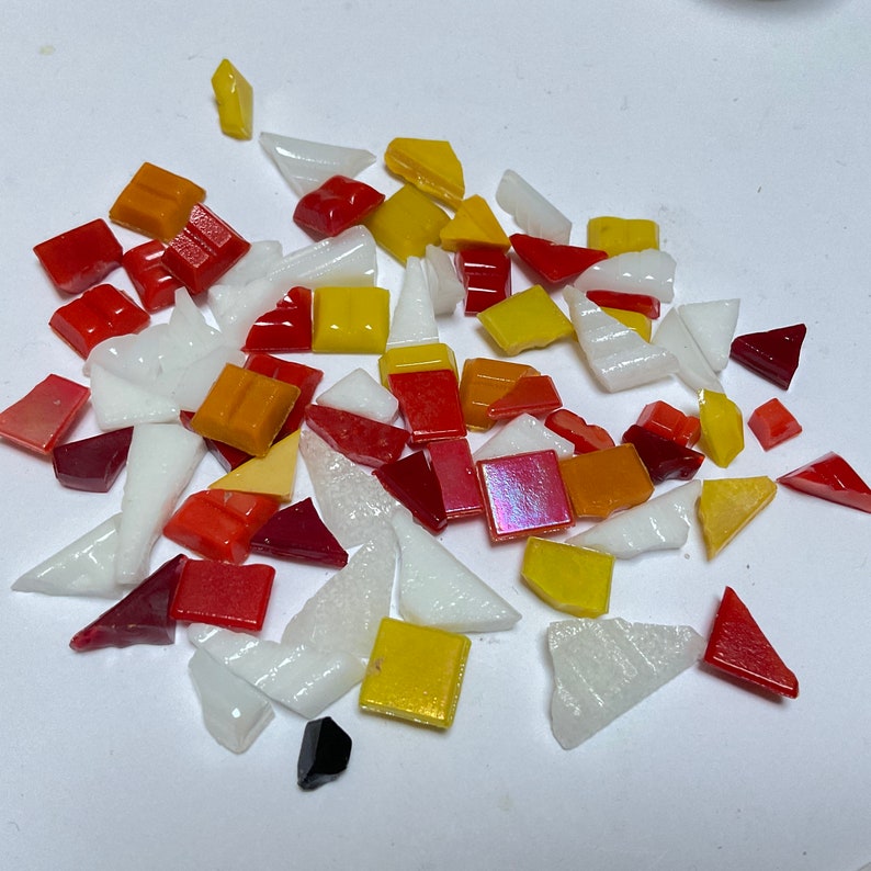 Puede incluir: Una colecci&oacute;n de coloridas teselas de mosaico de vidrio en varias formas y tama&ntilde;os. Las teselas son principalmente rojas, naranjas, amarillas y blancas, con algunas piezas iridiscentes y oscuras. Ideal para proyectos de arte de mosaico.