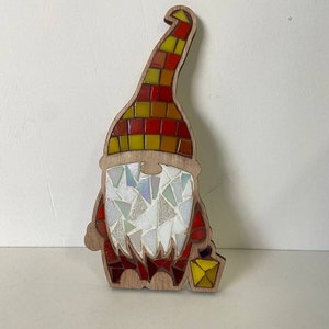 Puede incluir: Figura de gnomo de madera con dise&ntilde;o de mosaico. El sombrero del gnomo es un mosaico de cuadrados rojos, amarillos y naranjas. La barba est&aacute; hecha de piezas de mosaico blancas y transparentes iridiscentes, y el cuerpo es rojo. Una linterna amarilla est&aacute; en la mano del gnomo.