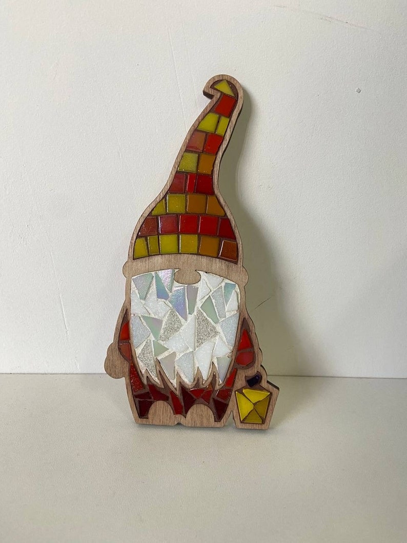 Puede incluir: Figura de gnomo de madera con dise&ntilde;o de mosaico. El sombrero del gnomo es un mosaico de cuadrados rojos, amarillos y naranjas. La barba est&aacute; hecha de piezas de mosaico blancas y transparentes iridiscentes, y el cuerpo es rojo. Una linterna amarilla est&aacute; en la mano del gnomo.