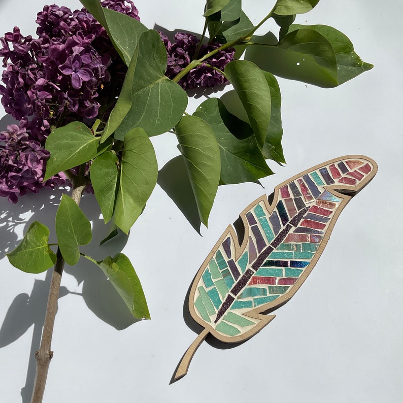 May include: A colorful mosaic feather made of small glass tiles in shades of blue, green, purple, and red. The feather is made of wood and has a natural wood grain.
