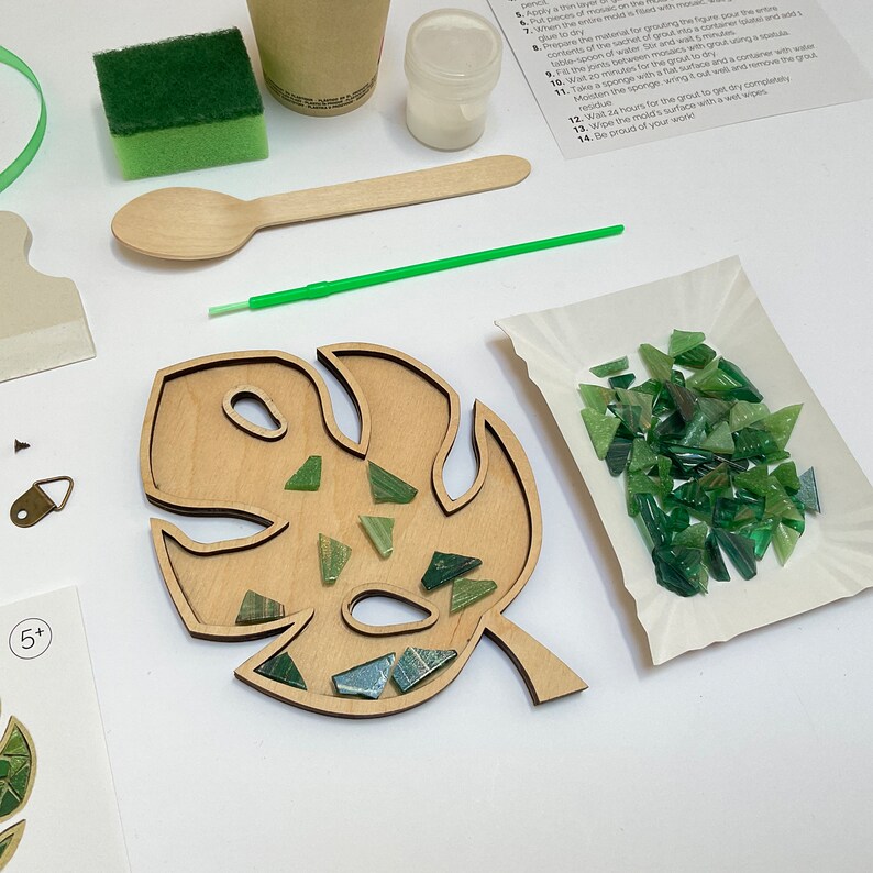 May include: A wooden monstera leaf shaped tray with a green and blue mosaic tile kit. The kit includes a small container of grout, a wooden spoon, a sponge, and a green plastic stick. The instructions are printed on a white sheet of paper.