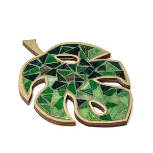 May include: A green mosaic leaf shaped like a monstera plant. The leaf is made of small, green glass tiles and is mounted on a wooden base.