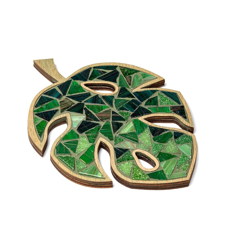 May include: A green mosaic leaf shaped like a monstera plant. The leaf is made of small, green glass tiles and is mounted on a wooden base.