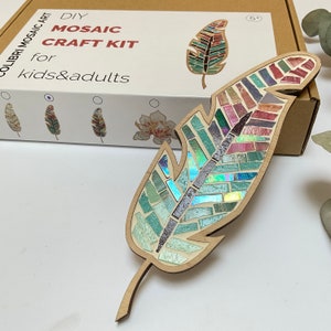 May include: DIY mosaic craft kit featuring a feather design. The kit includes a wooden feather shape with pre-applied mosaic tiles in shades of blue, green, pink, and purple. The box also shows other designs, including a flower and a leaf.