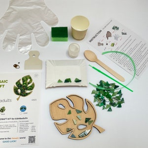 May include: A craft kit for making a mosaic art project. The kit includes a wooden leaf-shaped base, green glass mosaic tiles, glue, a sponge, and instructions. The kit is labeled "CRAFT KIT for kids & adults".