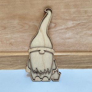 Puede incluir: Recorte de gnomo de madera clara con un sombrero largo y curvado, barba y linterna. El gnomo tiene contornos detallados. El objeto est&aacute; dise&ntilde;ado para ser un elemento decorativo.