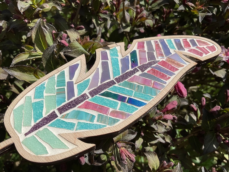 May include: A wooden feather mosaic with a colorful design. The feather is made of small pieces of glass in shades of blue, green, purple, and pink.