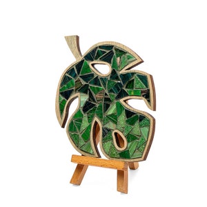 May include: A green mosaic leaf on a wooden easel. The leaf is made of small, green glass tiles and has a unique, organic shape.