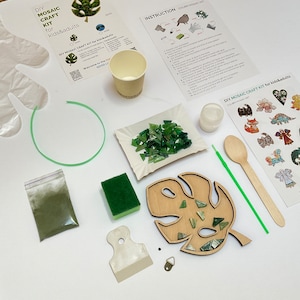 May include: DIY mosaic craft kit for kids and adults. The kit includes a wooden monstera leaf, green glass tiles, green glue powder, a wooden spreader, a green sponge, a small cup, a wooden spoon, a green straw, a small bag of green glitter, a metal hanging hook, and instructions.