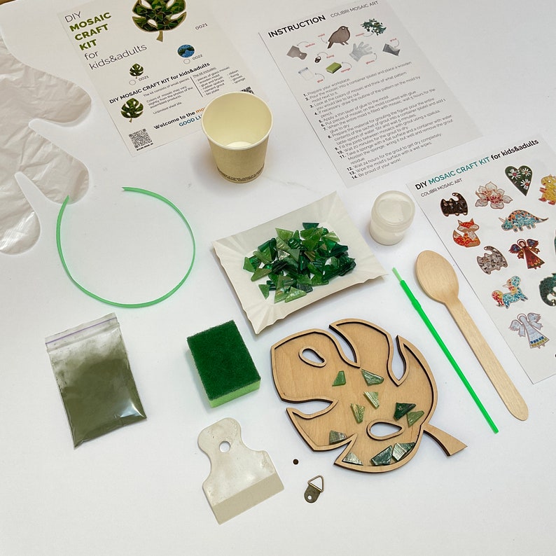 May include: DIY mosaic craft kit for kids and adults. The kit includes a wooden monstera leaf, green glass tiles, green glue powder, a wooden spreader, a green sponge, a small cup, a wooden spoon, a green straw, a small bag of green glitter, a metal hanging hook, and instructions.