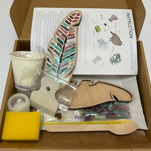May include: A craft kit for creating a mosaic art piece of a feather. The kit includes a wooden feather shape, a bag of colorful mosaic tiles, a small container of grout, a wooden spoon, a sponge, a scraper, and instructions.