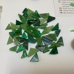 May include: A pile of green glass mosaic tiles in various shades of green. The tiles are triangular in shape and have a slightly iridescent finish.