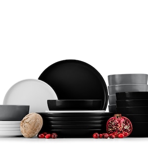 Op de afbeelding: Een set zwart, wit en grijs keramiek serviesgoed. De set bevat borden, kommen en een serveerschaal. Het serviesgoed is gerangschikt op een witte achtergrond met rode cranberries en een granaatappel.