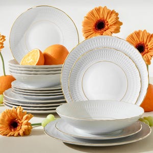 Könnte beinhalten: Weißes Geschirrset mit strukturiertem Muster und goldenem Rand. Das Set enthält Teller und Schalen, arrangiert mit Orangen und orangefarbenen Gerbera. Ein Stapel Teller und Schalen ist sichtbar.