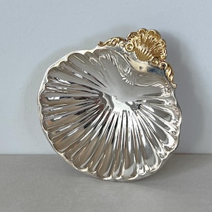 May include: A silver and gold shell-shaped dish with a decorative handle. The dish is made of metal and has a shiny finish.