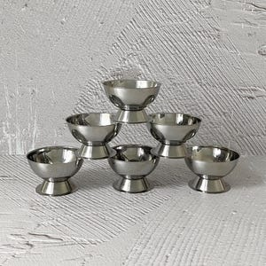 Könnte beinhalten: Sechs kleine Schalen aus Edelstahl sind pyramidenförmig angeordnet. Jede Schale hat eine abgerundete, becherartige Form auf einem Sockel. Die Schalen sind reflektierend, mit poliertem, metallischem Finish, vor einem strukturierten, hellgrauen Hintergrund.