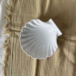 Könnte beinhalten: Weiße Keramik-Schale in Muschelform auf einem beigefarbenen Stoffhintergrund.