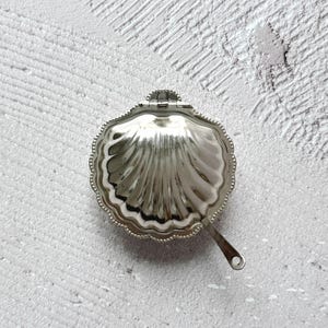 May include: A silver-coloured, seashell-shaped trinket box with a hinged lid and a small handle. The box has a scalloped edge and a textured, ridged interior. The handle has a small hole at the end.