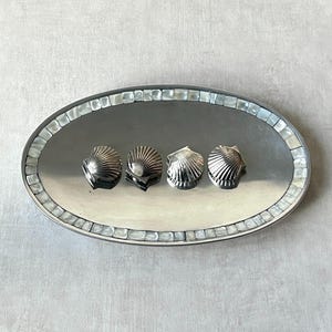 Puede incluir: Una bandeja ovalada plateada con un borde de mosaico de nácar. Cuatro objetos plateados en forma de concha marina están dispuestos en fila sobre la bandeja. La bandeja está sobre un fondo gris claro.