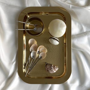 Può includere: Un vassoio rettangolare dorato con una trama martellata contiene vari oggetti a tema conchiglia. Questi includono una piccola ciotola con due utensili da portata, diverse conchiglie a forma di ventaglio e un ciondolo decorativo a conchiglia. Il vassoio è posto su uno sfondo bianco e serico.
