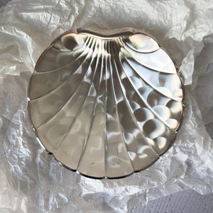 Può includere: Un piatto decorativo argentato a forma di conchiglia. Il piatto ha un bordo smerlato e una serie di linee curve e rigate che si irradiano dal centro. Il piatto è appoggiato su un letto di carta bianca stropicciata.